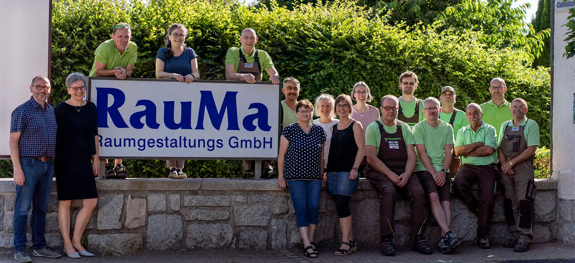 Die Gesamte Belegschaft der RauMa Raumgestaltungs GmbH um Lichtkasten mit RauMa Logo vor grüner Hecke